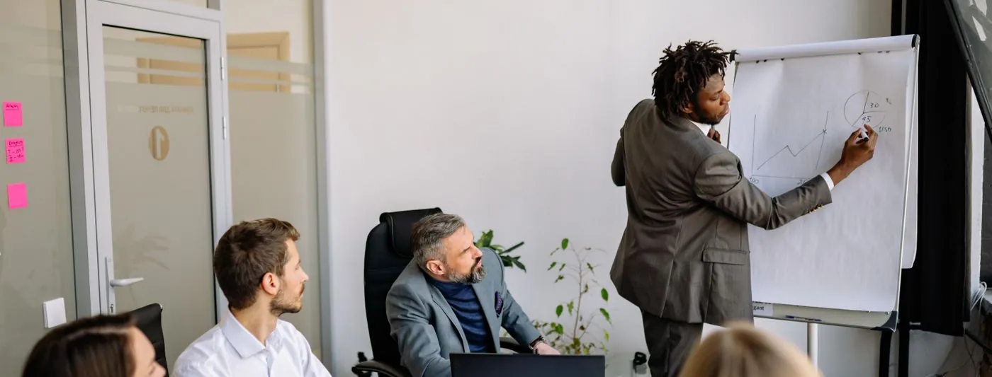 A facilitator writing on a board while colleagues discuss around a table.
