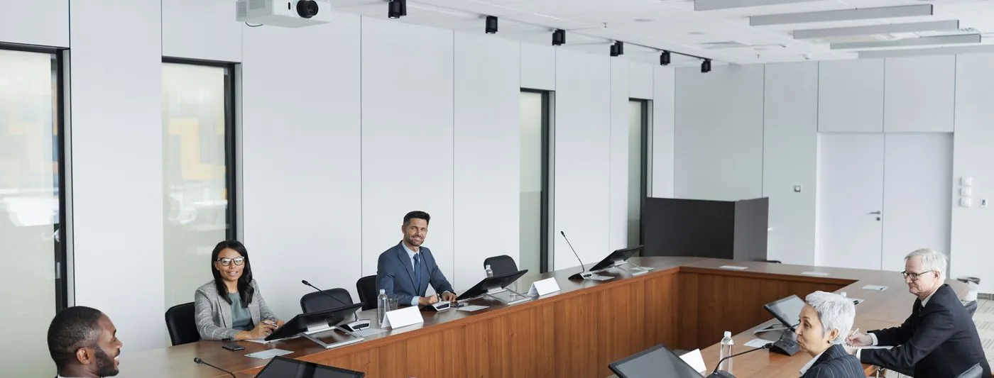 A wide institutional meeting room with participants seated around a long table.